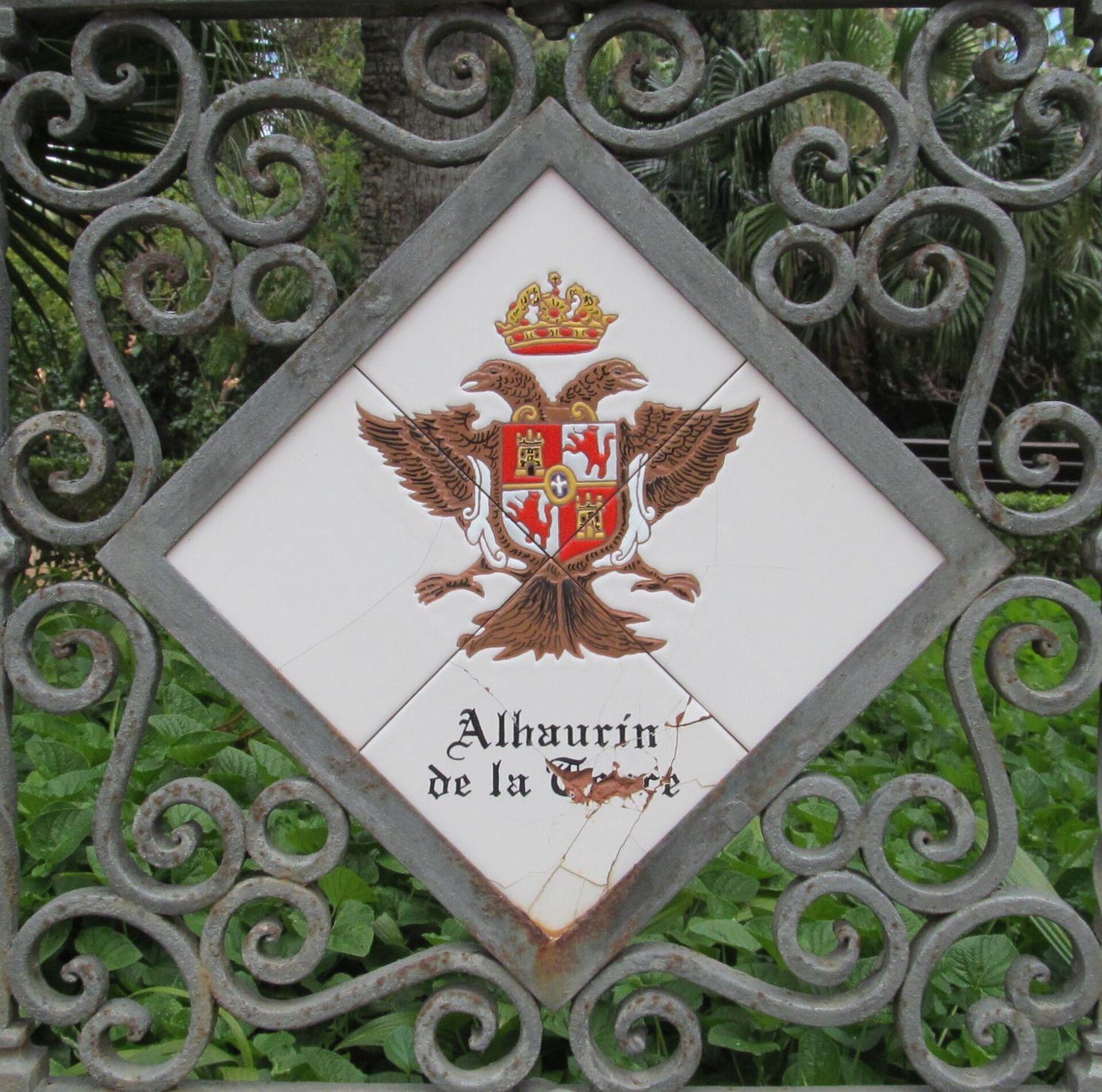 Diamond-shaped emblem with crown and eagle, surrounded by ornate metalwork, set in lush greenery.